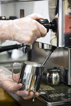A barista at a cafe churns milk in a metal jug using a caper of  professional coffee machine. Coffee house concept. The process of making cappuccino or latteの写真素材