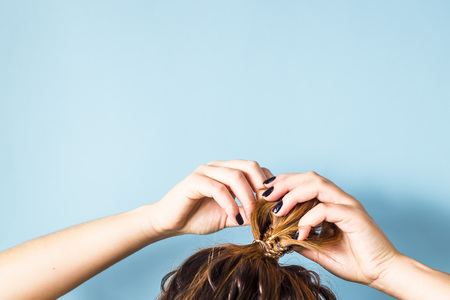 The woman straightens the disheveled bun on her head with her hands with a black manicure. Dark hair is tied with a transparent spiral elastic band. Modern fast hairstyle. Blue background. Copyspace