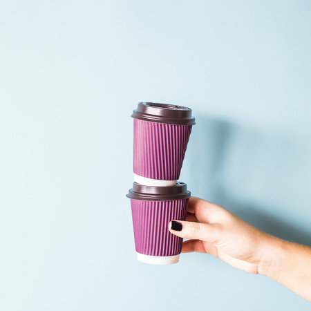 Purple paper kraft disposable cups for coffee with plastic lids in a female hand. One glass stands on the other. Tower of coffee. On blue backgroundの写真素材