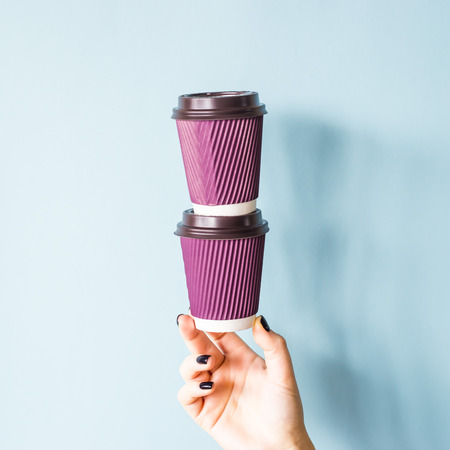 Purple paper kraft disposable cups for coffee with plastic lids in a female hand. One glass stands on the other. Tower of coffee. On blue backgroundの写真素材