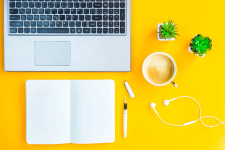 Workplace with a laptop, bkllonotom, pen, headphones, a cup of coffee and green whiskers in white pots. The working space of a freelancer, journalist, writer, modern worker. On a bright yellow background. Top view. Flat layの写真素材
