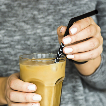 A girl in a gray dress adjusts a black straw in a large glass with ice-cold coffee with milk. Summer cooling drink conceptの写真素材