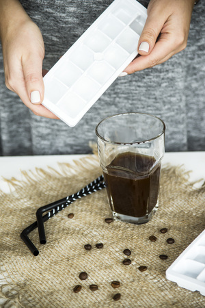 A girl in a gray dress is holding ice to add it to a glass of coffee. Preparation of a summer cooling drink conceptの写真素材