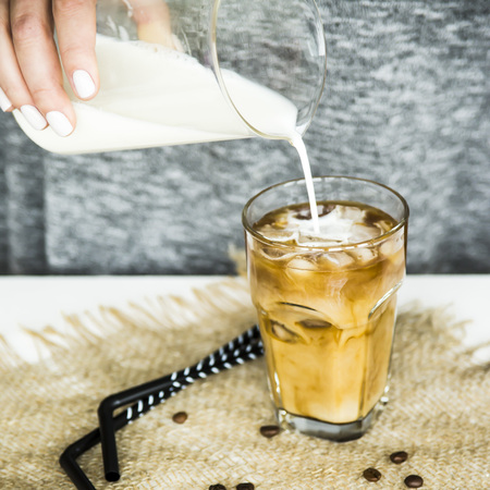 A girl in a gray dress pours milk into a large glass with ice coffee and a black straw. Preparation of a summer cooling drink conceptの写真素材