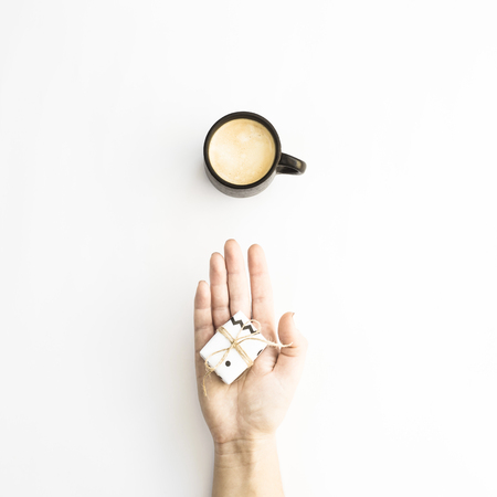 Female hands holding a small minimalist gift near a cup of coffee on a white background. Flat lay, top viewの写真素材