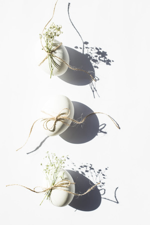 White minimalistic Easter eggs decorated with twine and flowers of gypsophila on a white background in bright light with deep shadows. Easter concept. Top view, flat layの写真素材