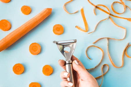 Carrot cleaning with a special kitchen appliance in female hand. On a blue background. Bright carrot shavings and rings. Top view, flat layの写真素材