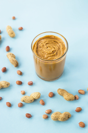 A glass jar with peanut butter paste among the peanuts in shell and without on a blue background. Vegan product. American Breakfastの写真素材