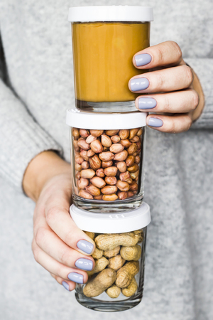 The woman holds in her hands three jars - with peanuts in a shell, peeled peanuts and peanut butter. Vegan and natural food conceptの写真素材