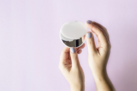 A compact, compact mirror in women's hands. On a lilac bright background. Makeup female accessoriesの写真素材