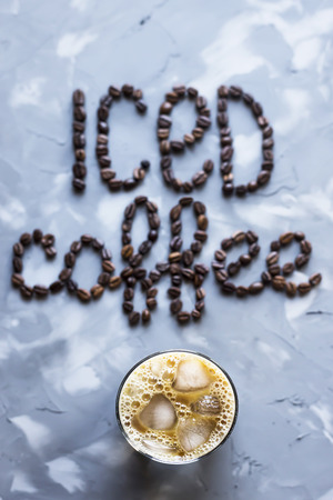 A glass of iced coffee with milk next to the inscription "ice coffee" from coffee beans on a gray concrete background. Summer cooling drinkの写真素材