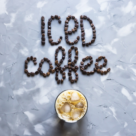 A glass of iced coffee with milk next to the inscription "ice coffee" from coffee beans on a gray concrete background. Summer cooling drinkの写真素材