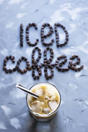 A glass of iced coffee with milk next to the inscription "ice coffee" from coffee beans on a gray concrete background. Summer cooling drinkの写真素材