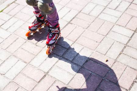 A little girl rolls on red rollers on the sidewalk. Shadow on the road. Active rest conceptの写真素材