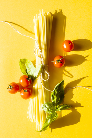Dry spaghetti on a yellow background next to cherry tomatoes and a basil leaf. Top view, flat layの写真素材