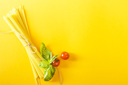 Dry spaghetti on a yellow background next to cherry tomatoes and a basil leaf. Top view; flat layの写真素材