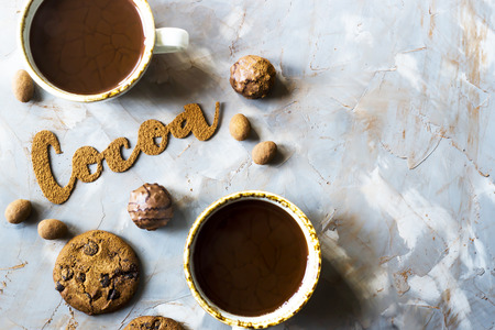 The word cocoa is written in letters of cocoa on a gray concrete background next to two cups of cocoa and chocolate sweets and biscuits. I love the cocoa conceptの写真素材