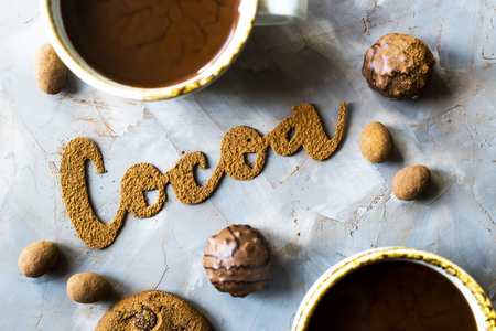 The word cocoa is written in letters of cocoa on a gray concrete background next to two cups of cocoa and chocolate sweets and biscuits. I love the cocoa conceptの写真素材
