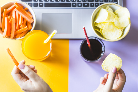 A woman chooses between a harmful and healthy snack. Chips and cola or carrots and juice. Eating in a break between working on a laptop. Junk Food Conceptの写真素材