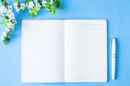 An open blank notebook with a white pen and blossoming branch on a blue background. Spring workplace conceptの写真素材