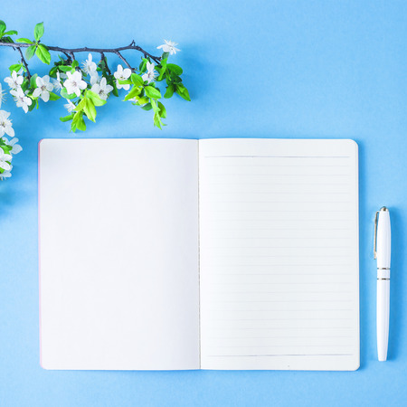 An open blank notebook with a white pen and blossoming branch on a blue background. Spring workplace conceptの写真素材