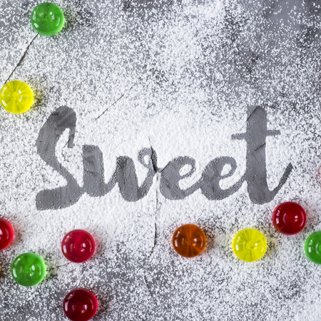 The word sweet is written on a gray concrete background with the help of powdered sugar next to colorful candy. Top view, flat layの写真素材