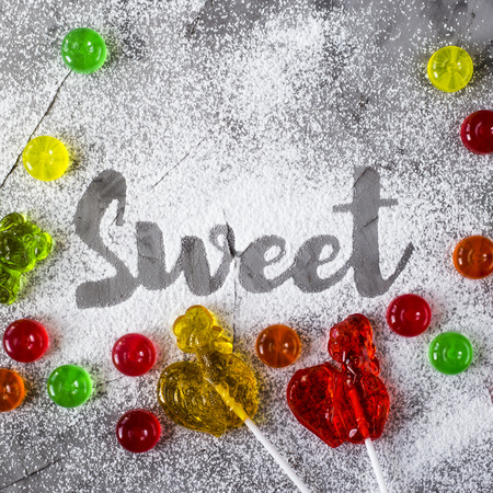The word sweet is written on a gray concrete background with the help of powdered sugar next to multi-colored sweets and retro candies in the form of cockerels. Top view, flat layの写真素材