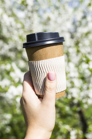 A woman's hand holds a disposable cup of coffee against the blossoming spring tree. The beginning of a beautiful spring day. Good mood and lifestyleの写真素材