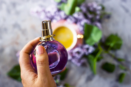 A female hand holds a purple perfume bottle against the background of a cup of coffee and lilac. Beauty and fragrance for women conceptの写真素材