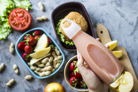 Cooking lunch for the child to school. On the gray kitchen table. Sandwich, strawberries and peanuts in lunchboxes. Top view. A woman holding a bottle of juiceの写真素材