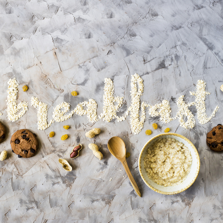 The word breakfast is written on a gray background of oatmeal. Next to a bowl of ready-made oatmeal, spoon, nuts, raisins and chocolate biscuits. Minimalism. Top view, flat layの写真素材