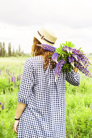 Caucasian girl in a blue dress with a hat is holding a bouquet of blue wildflowers of lupines in her hands. Recreation in the spring on a nature outside the city conceptの写真素材