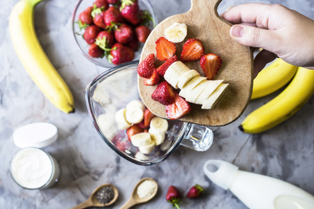 Ingredients for making strawberry banana smoothies on a gray table next to a glass bowl of blendet. Cooking a healthy breakfast concept. TOp view, flat layの写真素材