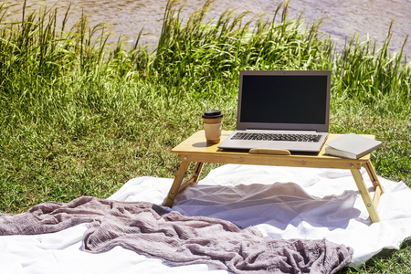 Freelancer work in summer on a picnic in nature. Laptop on a bamboo table on a coverlet by the riverの写真素材