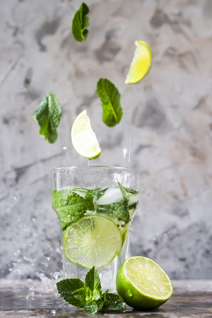 Leaves of mint and lime fly into a glass with mojito with ice on a gray background. Levitation flying magic items. Summer drink. With a spray of waterの写真素材