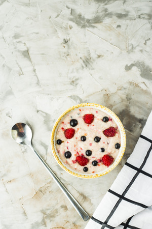 A bowl of oatmeal with raspberries and blueberries on a gray table next to a white checkered napkin. Healthy summer breakfast. Top view, flat lay, copyspaceの写真素材