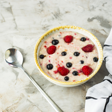 A bowl of oatmeal with raspberries and blueberries on a gray table next to a white checkered napkin. Healthy summer breakfastの写真素材
