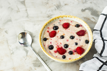 A bowl of oatmeal with raspberries and blueberries on a gray table next to a white checkered napkin. Healthy summer breakfastの写真素材
