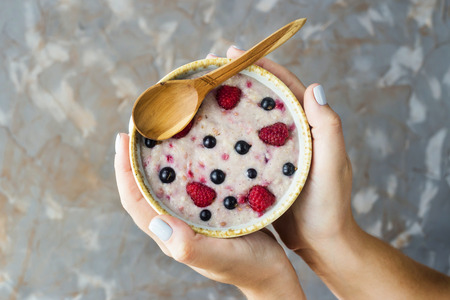 A bowl of oatmeal with raspberries and blueberries in female hands on a gray background. Healthy summer breakfast conceptの写真素材
