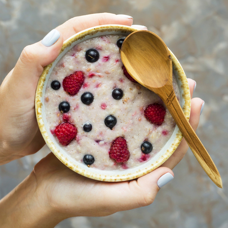 A bowl of oatmeal with raspberries and blueberries in female hands on a gray background. Healthy summer breakfast conceptの写真素材