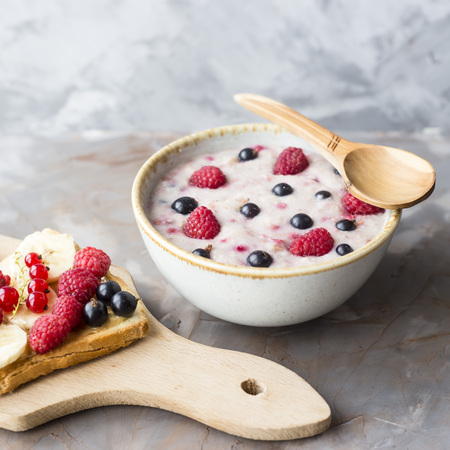 A bowl of oatmeal with berries next to a board with sandwiches with peanut butter and banana, sporodina, raspberries. Healthy summer breakfast on a gray tableの写真素材