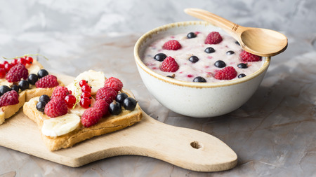 A bowl of oatmeal with berries next to a board with sandwiches with peanut butter and banana, sporodina, raspberries. Healthy summer breakfast on a gray tableの写真素材