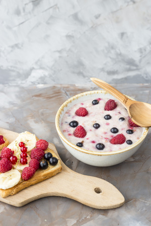 A bowl of oatmeal with berries next to a board with sandwiches with peanut butter and banana, sporodina, raspberries. Healthy summer breakfast on a gray tableの写真素材
