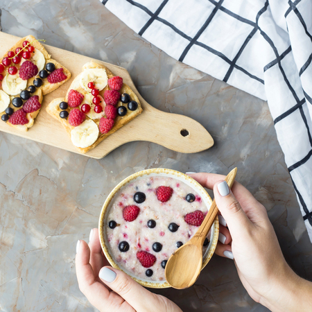 Oatmeal with berries and sandwiches with peanut butter, banana, currant and raspberries on a gray table with female hands. Top view, flat layの写真素材