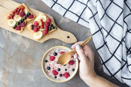 Oatmeal with berries and sandwiches with peanut butter, banana, currant and raspberries on a gray table with female hands. Top view, flat layの写真素材
