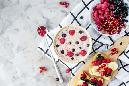 A table with a healthy summer breakfast - oatmeal with berries, currants and raspberries, and a sandwich with peanut butter, banana and various berries. Top view, flat layの写真素材