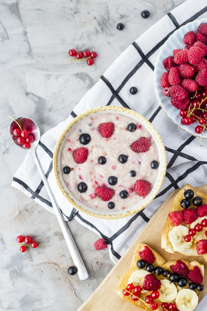 A table with a healthy summer breakfast - oatmeal with berries, currants and raspberries, and a sandwich with peanut butter, banana and various berries. Top view, flat layの写真素材