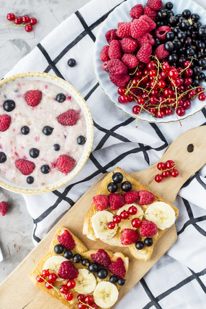 A table with a healthy summer breakfast - oatmeal with berries, currants and raspberries, and a sandwich with peanut butter, banana and various berriesの写真素材