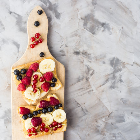 Sendvci with peanut butter, banana and raspberries and currants on a wooden board on a gray table. Vegan healthy breakfast. Top view, flat lay, copyspaceの写真素材