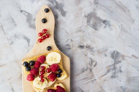 Sendvci with peanut butter, banana and raspberries and currants on a wooden board on a gray table. Vegan healthy breakfast. Top view, flat lay, copyspaceの写真素材